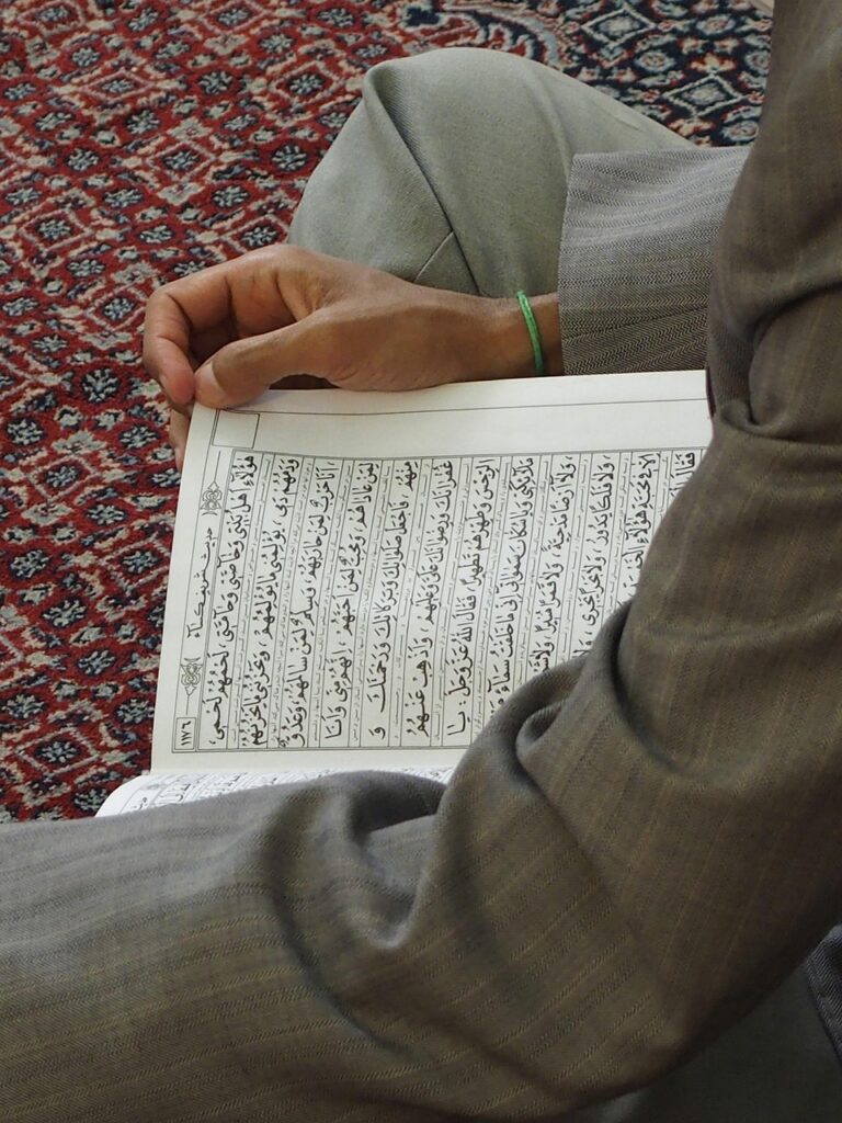 Homme assis sur un tapis de prière lisant une page du Coran ouverte, illustrant l’apprentissage et la récitation au quotidien
